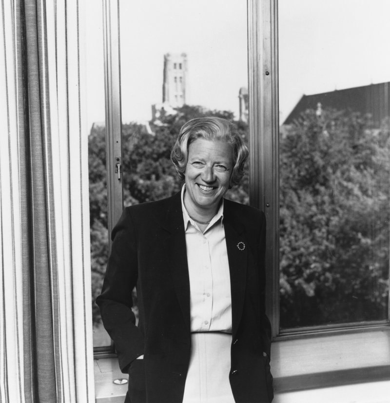 Photo of Hanna Holborn Gray in 1981 standing in front of a window with Rockefeller tower in the distance. She is smiling and wearing a dark blazer and has her hands in her pockets.