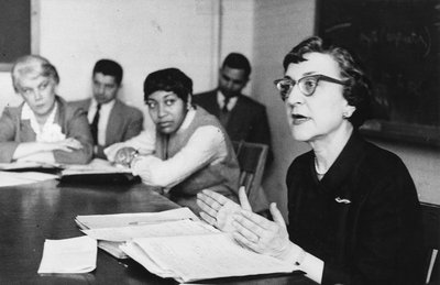 Black-and-white photograph of Helen Harris Perlman, the Distinguished Service Professor in the School of Social Service Administration at the University of Chicago, meeting with an unidentified group. Helen is a middle-aged white woman with dark, permed hair. She is wearing a dark dress and glasses. She is seated in the right foreground of the photograph at a large wooden table. She has papers on the table in front of her and is gesturing with both hands as she speaks. In the background, two women and two men are seated at or near the same table and are listening to Helen speak. Clothing and hairstyles of the people in the photograph suggest that the photograph dates to the 1960s.