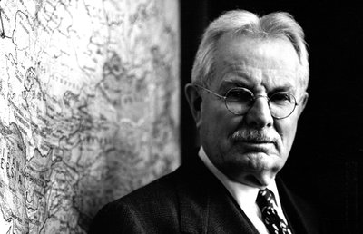 Black-and-white photograph of Quincy Wright, Professor of International Law at the University of Chicago. Wright is shown from the chest up, standing in front of a world map. He is looking directly into the camera and is wearing a dark suit and tie and glasses. He is a middle-aged white man with white hair and a mustache.