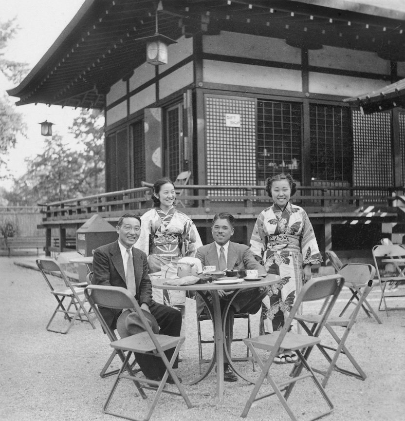 Two men sit at a cafe table in the Japanese Garden, Jackson Park. Two women in kimonos stand behind them.