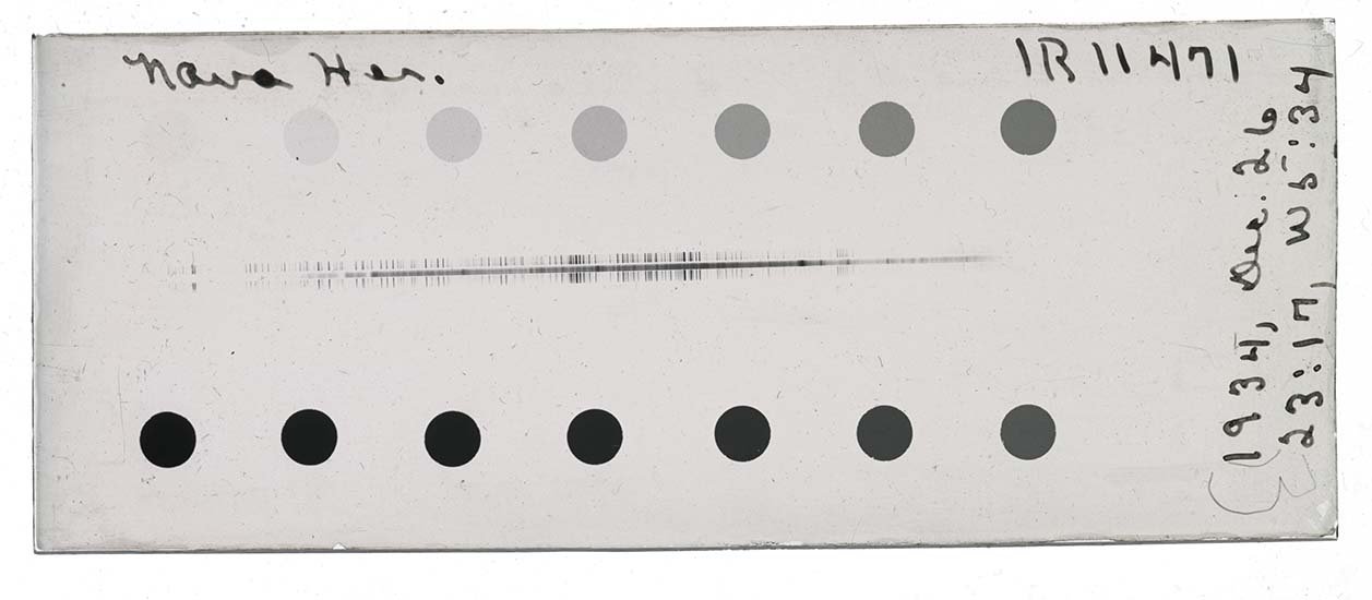 A glass plate negative that is about 2x wider than it is tall. In the middle, the spectra is a series of dark vertical lines of varying heights and densities. It is nearly horizontal, but raises a bit on the right. Above and below the spectra are a series of circles that increase in darkness from left to right in a clockwise direction. The lightest is on the top left, the darkest is on the bottom left. The caption on the slide is handwritten in ink and says "Nova Her. 1R11471" across the top. On the right edge it reads, "1934, Dec. 26" and below "23:17, W5:34".