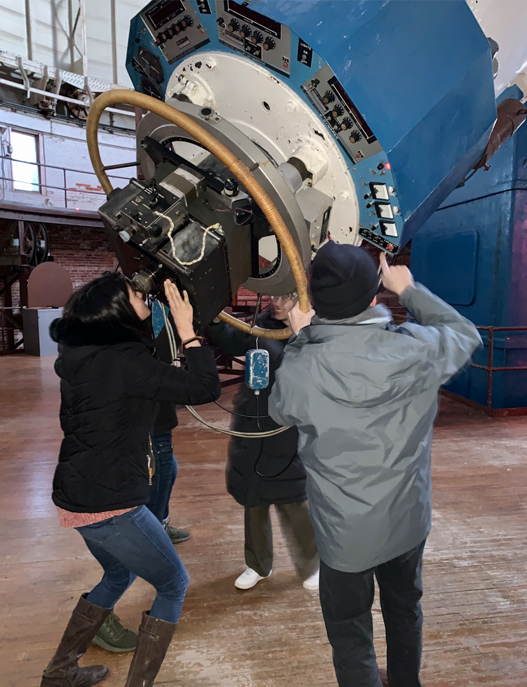 A woman in a black jacket and blue jeans bends her knees to look through the eyepiece of a large, blue telescope.  A man in a gray coat wearing a black beanie hat stands to her right. He has one hand on the telescope and is pointing to it with the other. There is a person in the background whose legs show behind the telescope.