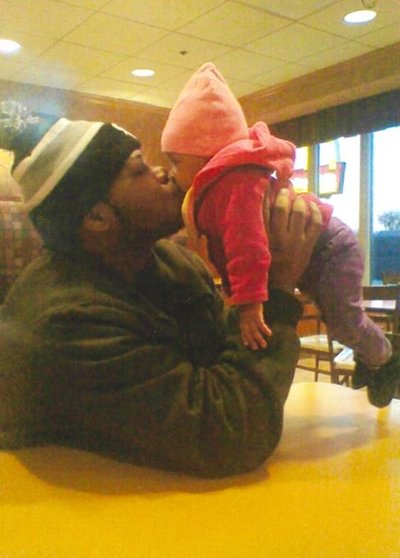 A twenty something, dark skinned Black man sitting in a booth at a restaurant. He is wearing a knit hat with a pompom on top and a black, zipped up coat. He is holding an infant in his hands up to his face to give them a kiss.