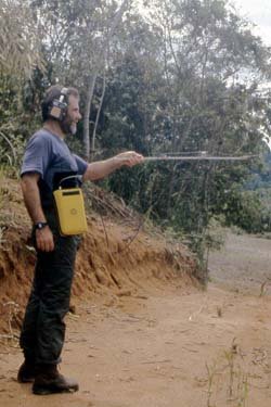 A man stands on a dirt path with a metal-detector-like object.