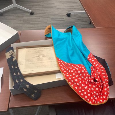 large, flat gray archival box open on a brown table. An framed, honorary degree is inside with a sock and an academic rob laid over the sides.