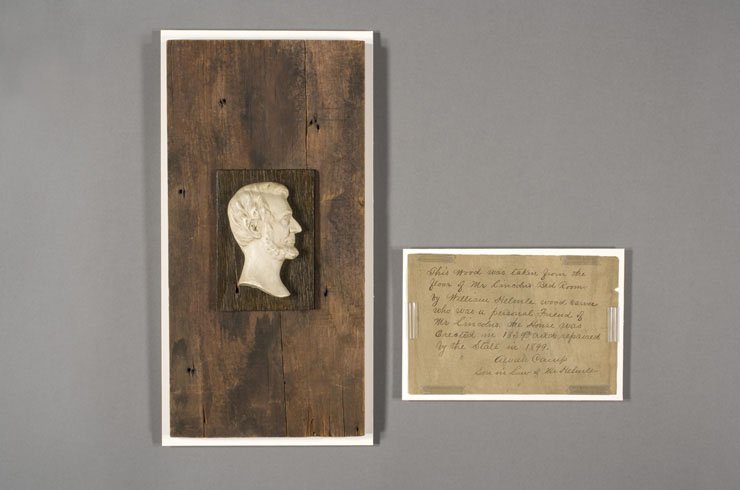 A dark wooden board below a white carving of Lincoln's bust, alongside an old postcard filled with script.
