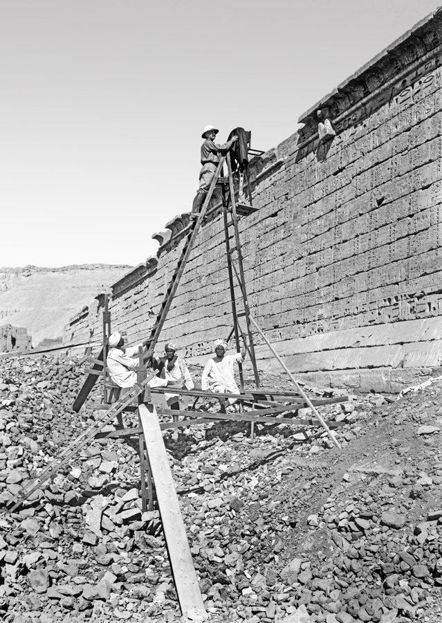 Man at top of ladder and three men near base of ladder near wall