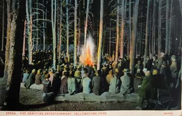 A campfire in a forest with many people sitting around.