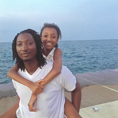 A dark skinner, twenty something Black man with a thin moustache and goatee is standing in front of Lake Michigan. He has shoulder length locks and is wearing a white T-shirt. He is holding a light skinned Black child on his back, whose arms are around his shoulders. Both are smiling.