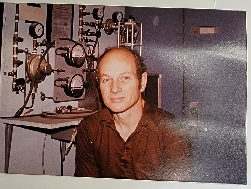 Color photograph of James Cronin seated, wearing brown button-down shirt with lab instruments behind him.