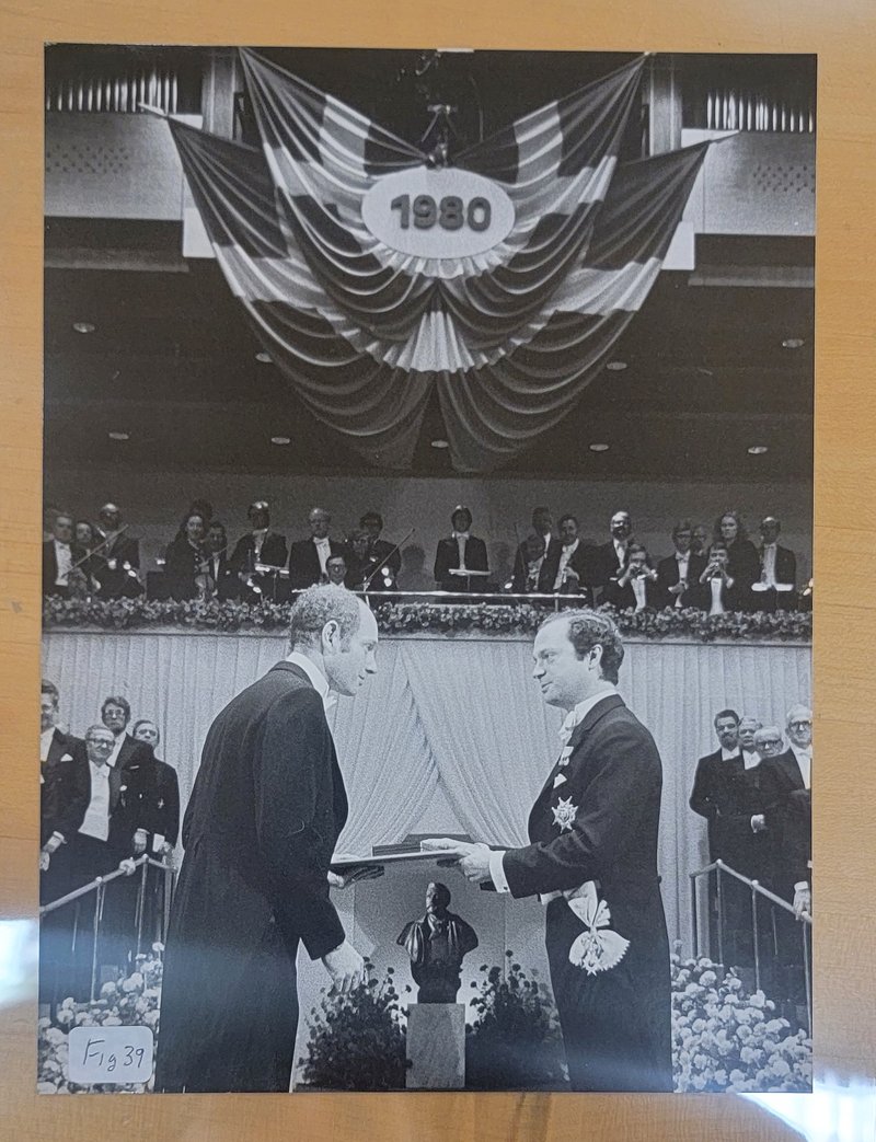 Black-and-white photo of James Cronin in tuxedo accepting the Nobel Prize from HM King Carl XVI Gustaf of Sweden. Bunting with the year 1980 is displayed above them, and audience members are visible in the background.