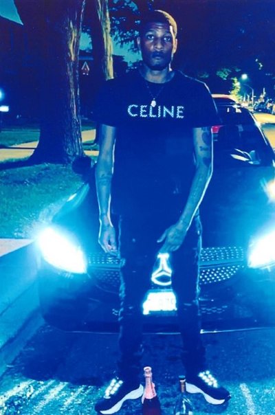 A medium dark skinned Black man with closely shorn hair stands in front of a black BMW with its headlight on. He is looking at the camera. It is twilight and you can see houses and trees in the background. He is wearing a jeans and a black T-Shirt with "Celine" printed on the front. His left hand covers his groin and there is an open champagne bottle at his feet.