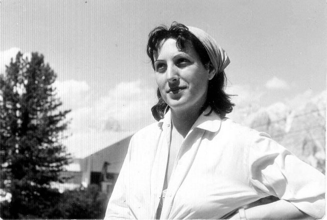 A black and white portrait of a woman in a white shirt standing outside. There are mountains and a tree in the background.