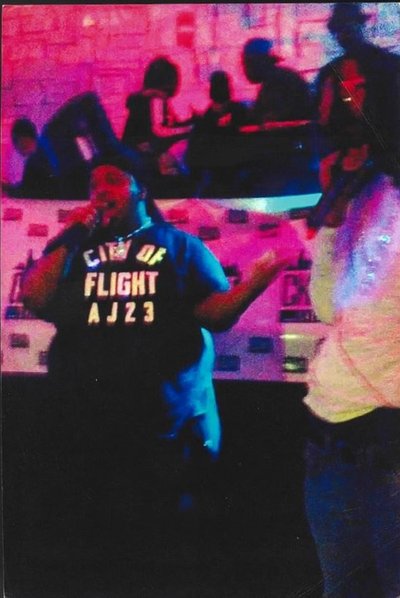 A Black man with medium dark skin, stands in a restaurant in front of a banner. He is speaking into a microphone in his hand. He wears a black durag on his head and a black t-shirt with white lettering "City of Flight AJ23," and a silver necklace. There is a person with a microphone, standing to his left. There are people sitting at tables behind him. There is pink lighting in the room.