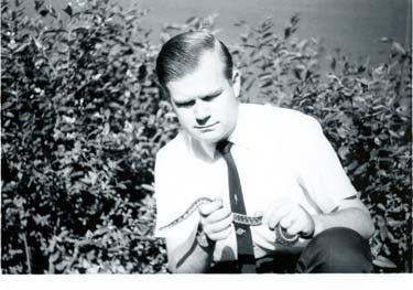 A man in a suit sits in a bush, holding a small snake.