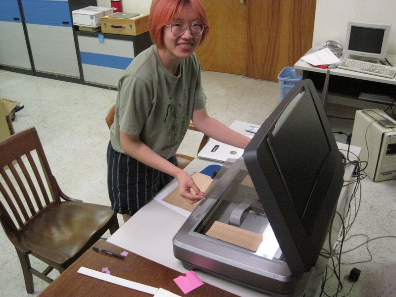 A person with chin-length reddish hair, glasses, a green shirt and striped pants is smiling as they hold a glass plate over an open, tabletop scanner.