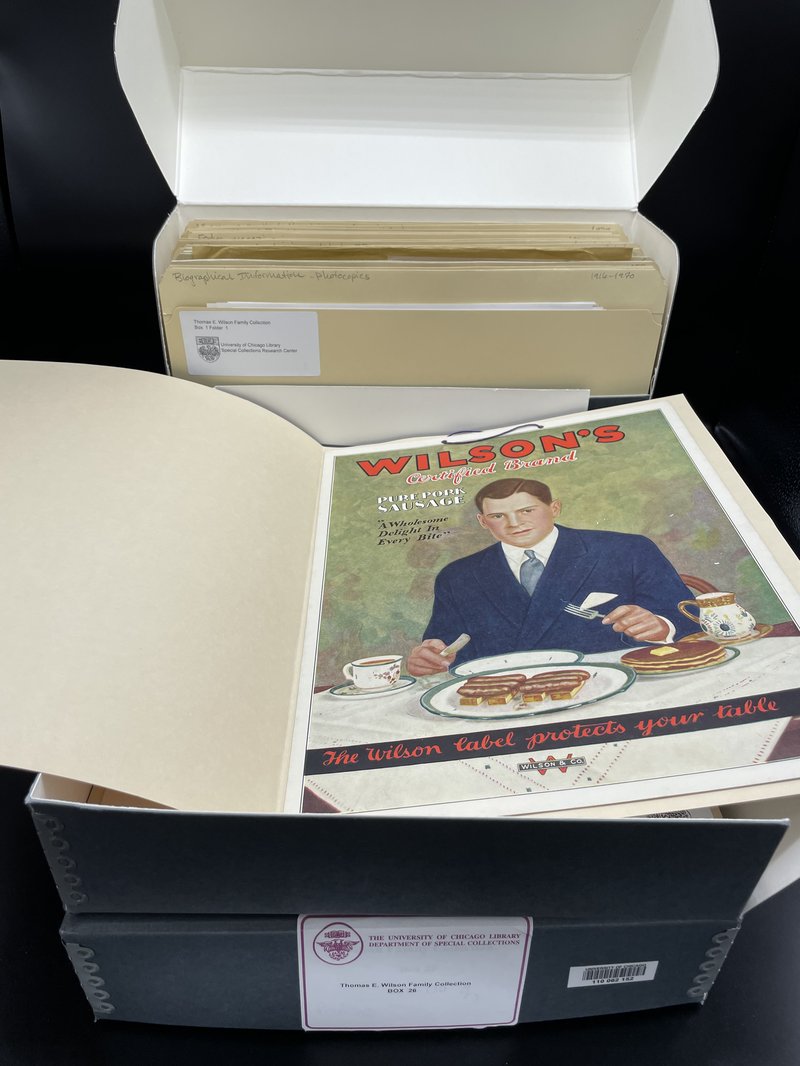 Photograph of three archival boxes from the Thomas E. Wilson Family Collection. Two boxes are open to reveal folders inside. One folder is open, showing a 1920s Wilson & Co. print advertisement for pork sausage, featuring Edward Foss Wilson seated at a breakfast table.