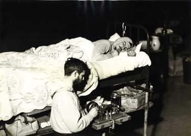A man fiddles with medical equipment next to a bedridden man asleep.