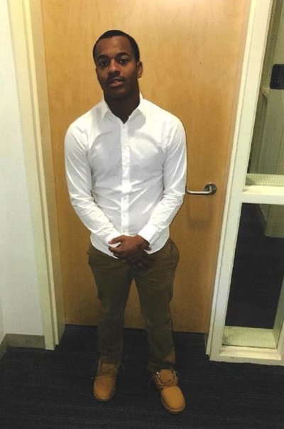 A Black man with short hair in his 30s wearing a white button down shirt, brown pants, and brown hiking boots, standing in front of a door to an office