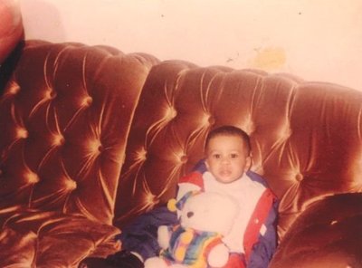 Jonathan Hurst as a baby sitting on a brown, velvet couch, holding a stuffed bear and looking at the camera