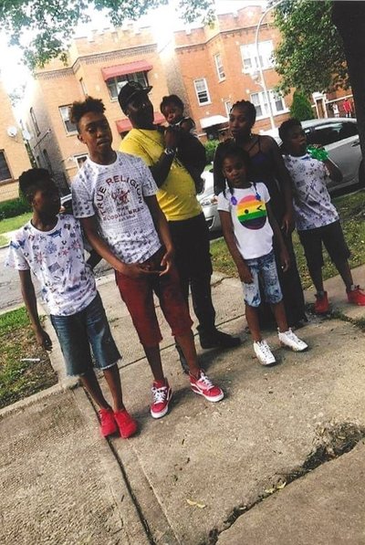 A family of 7, two adults and 5 children, standing on the sidewalk in summer. Aaron Moore is in the middle, holding a baby. He is a Black man in his forties, wearing a yellow T-shirt, a black cap and black jeans.