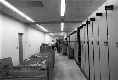 Cardboard boxes full of books next to rows of bookshelves.