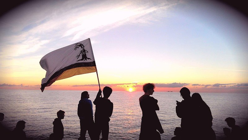 A man waves  flag with a smiling tree on it in front of Lake Michigan as the sun rises.
