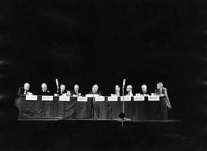 About ten men with namecards sit onstage behind a table.