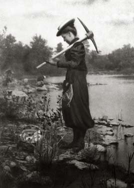A woman in old dress stands in a swamp, carrying a pickax.