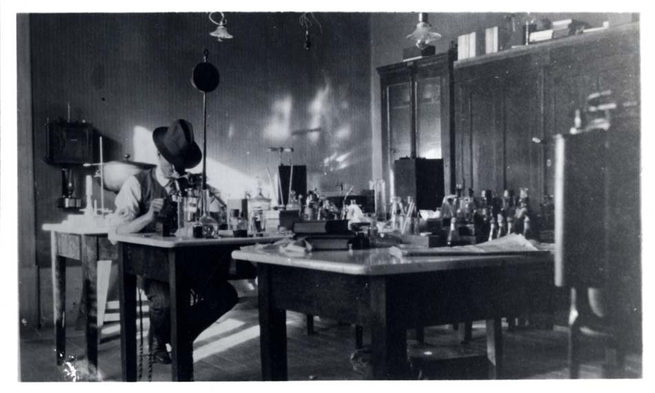 Man seated in lab looking into a microscope. He has a fedora on and is wearing it tilted forward.