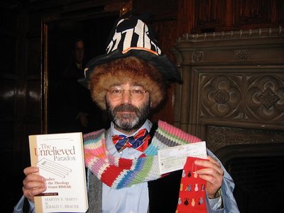 A 50 something white man with black hair and a beard, holding a book and check wearing a silly hat and scarf