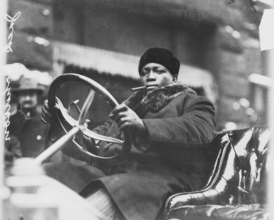 Jack Johnson sitting in an open car holding a steering wheel.