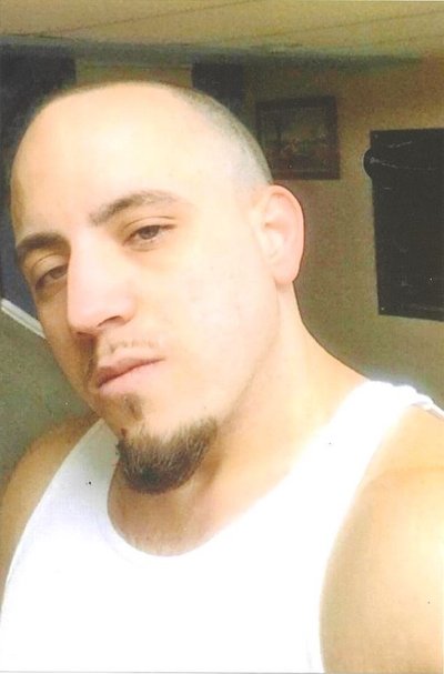 Selfie of a man with light tone skin in their thirties. He has a shaved head and goatee. He is wearing a white sleeveless shirt. You can see pictures hanging on the wall behind him.