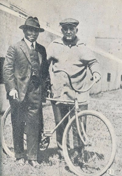 A black and white photograph of Stagg and Okabe. They are both standing, and Stagg is holding a bicycle.