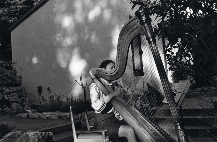 Photograph of a woman playing a harp in a garden.