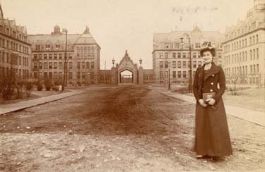 An old-fashioned photo of a woman in a field surrounded by Gothic buildings.