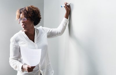 Color photograph of African American woman standing in front of a white board. She is holding a piece of paper in one hand, and a dry erase marker in the other, poised to write on the white board. She is partially turned away from the white board and is speaking. This is a linked image.