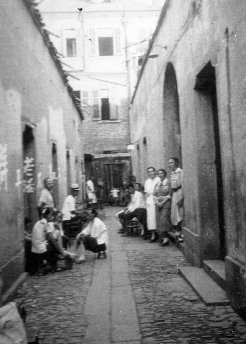 An old photo of men and women standing together in a cobblestone alley, surrounded by buildings with Chinese characters.