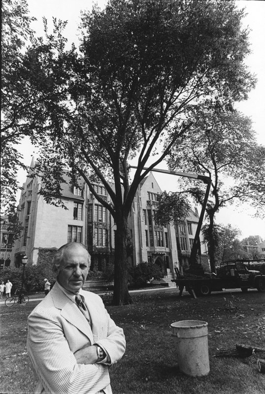 A black and white photo of Herbert L. Anderson standing in front of a large tree in front of a university building. In the background is a plastic garbage can and a crane where someone is trimming the tree. Anderson wears a striped suit jacket and a tie, and his face is in a grimace.