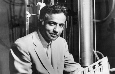 Black-and-white photograph of Subrahmanyan Chandrasekhar, the Morton D. Hull Distinguished Service Professor in the departments of Astronomy and Astrophysics at the University of Chicago, and recipient of the Nobel Prize in Physics for 1983. Chandrasekhar is shown seated in front of laboratory equipment. He is wearing a light-colored suit, and is smiling slightly into the camera. He holds an open book on his lap. Chandrasekhar is a middle-aged Indian-American man.