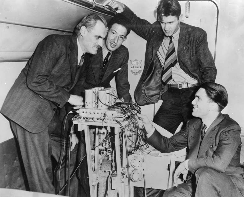 A black and white photo of four men in suits gathered around a piece of equipment