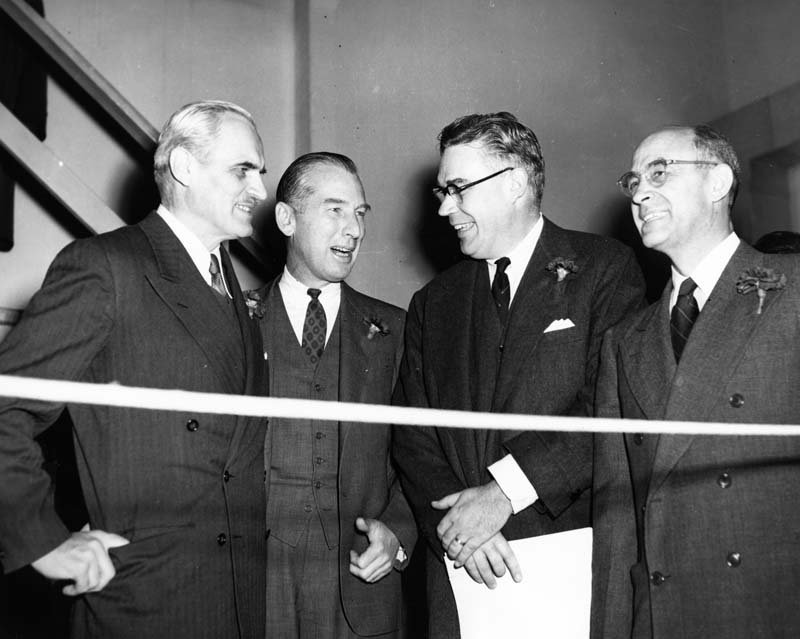 Four men wearing black suits and ties stand in a semi-circle, smiling and talking to each other.