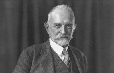 Black-and-white photograph of George Herbert Mead, philosopher and sociologist. Mead is an older white man with short white hair and a white mustache and beard. He is wearing a dark suit, vest, and tie. He is looking directly into the camera. Behind him is a dark curtain.