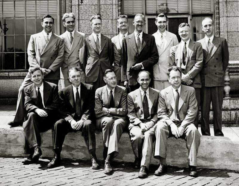A black and white photo of thirteen men in two rows. They all wear suits and ties and are looking off to the right of the photo.