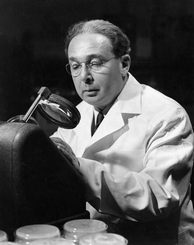 A black and white photo of Leo Szilard looking through a magnifying glass in a white lab coat.