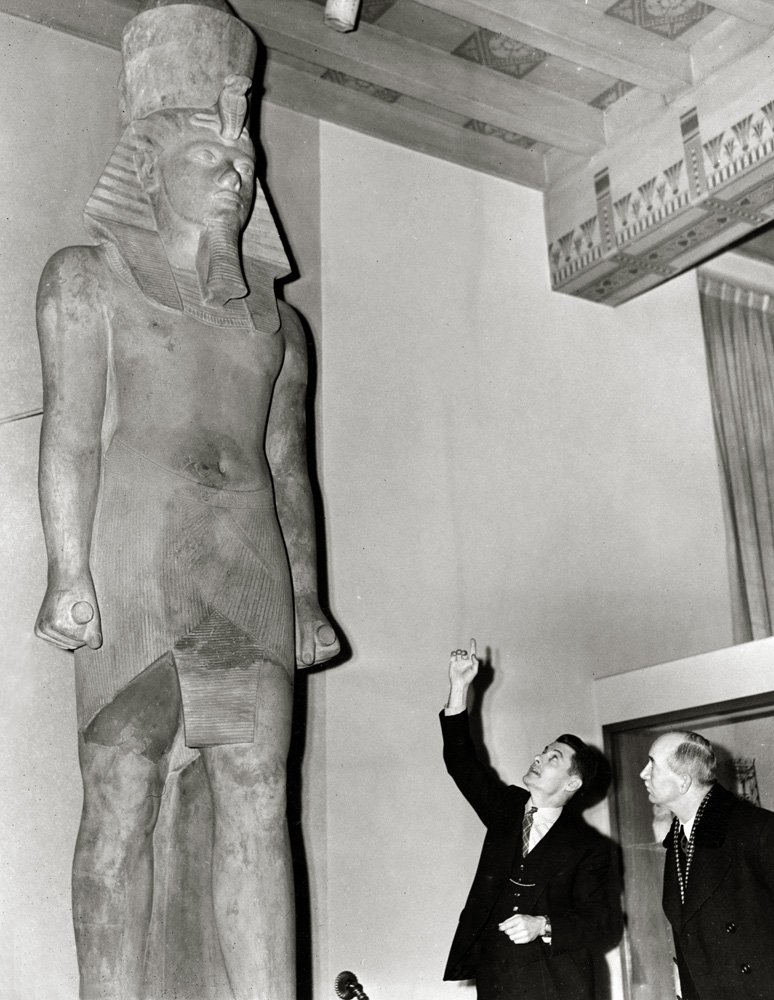 Two men gesture towards the Colossal Statue of Tutankhamun.