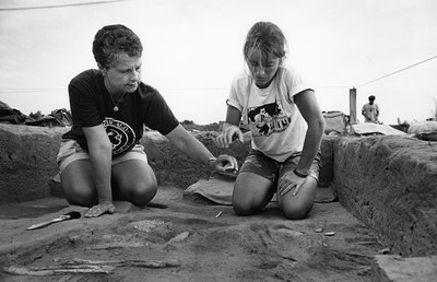 Black-and-white photograph of Jane E. Buikstra, bioarchaeologist and professor of Anthropology at the University of Chicago. She is pictured with a student at a Hopewell excavation site. Buikstra is a middle-aged white woman with short, dark, curly hairy. She is wearing a UChicago t-shirt and shorts, and is kneeling in an excavation site next to the student. The student is a young, white woman with blonde hair. She is wearing her hair in a ponytail, and is wearing a white graphic t-shirt and shorts. Both women are looking down at a small object that Buikstra is holding out in her left hand.