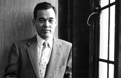 Black-and-white photograph of Joseph M. Kitagawa, dean of the Divinity School and professor in the department of East Asian Languages and Civilizations at the University of Chicago. Kitagawa is shown standing, leaning his left shoulder against a window. He is wearing a light-colored suit and tie. Kitagawa is a young, Japanese American man. He is looking directly into the camera with a slight smile.