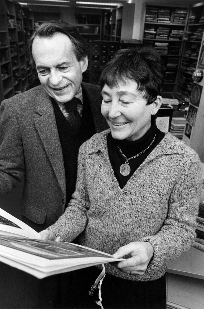 Black-and-white photograph of Lloyd and Susanne Rudolph. Lloyd is a white man with dark hair, wearing a tweed jacket, shirt, tie, and sweater vest. Susanne is a white woman with short, dark hair, wearing a knit sweater over a dark turtleneck shirt  and a necklace with a large pendant. The couple are standing together looking at a folio-size bound image. Lloyd stands behind Susanne and looks over her shoulder. Susanne is holding the folio. They are both smiling. Behind them, bookshelves and filing cabinets are visible.