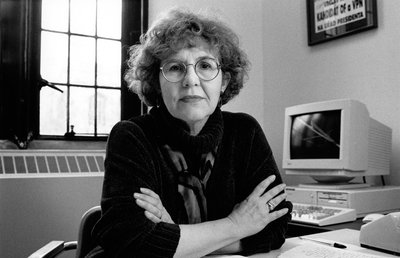 Black-and-white photograph of Jean Bethke Elshtain,  the Laura Spelman Rockefeller Professor in the Divinity School, the Department of Political Science and the Committee on International Relations at the University of Chicago, seated at desk in office at the University of Chicago. Elshtain is a middle-aged white woman with short curly hair. She is wearing glasses, and a dark sweater and scarf. She is looking directly into the camera with a serious expression, and has her arms crossed with elbows resting on the desk. Behind her is a 1990s PC and a window.